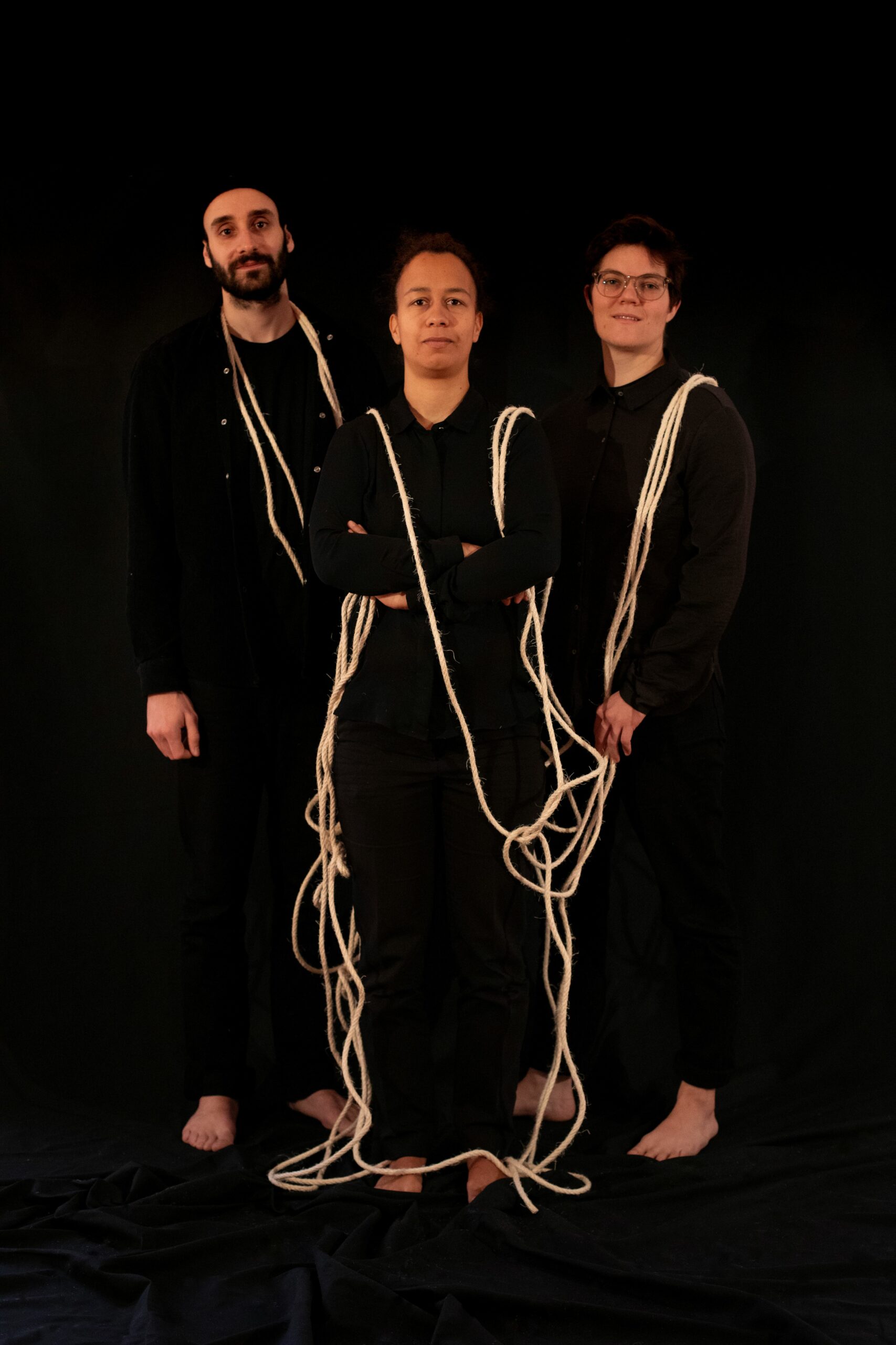 Trio Kokon stehend, in Schwarz gekleidet, helle Schnüre verbinden sie